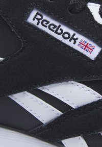 Zapatilla negra con una superficie texturizada, detalles en cuero blanco y un logo de Reebok con una pequeña bandera del Reino Unido. Diseñada para la comodidad y flexibilidad.