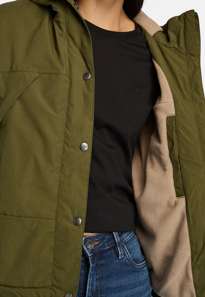 Giacca verde oliva con cappuccio e bottoni a pressione metallici, indossata sopra una t-shirt nera con un logo ricamato, dotata di un rivestimento beige.