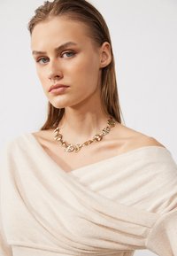 Off-white draperad topp med vid rundhalsring, med subtil textur. Halsband i guldton med länkade blommoliknande former vilar på nyckelbenet.