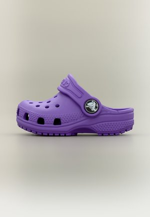 Sabot en caoutchouc violet avec des trous de ventilation et une bride arrière réglable, arborant un badge logo crocodile noir et blanc sur un fond neutre.