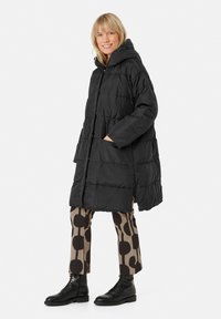 Giacca a vento nera con cappuccio, design trapuntato, tasche laterali e maniche lunghe, abbinata a pantaloni beige e neri con motivo e stivali neri.