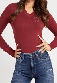 Haut bordeaux à manches longues avec encolure en V, texture côtelée et accents en strass, associé à un jean en denim bleu taille haute avec des détails usés.