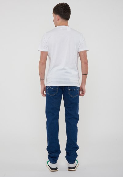 T-shirt bianco a maniche corte e jeans blu a gamba dritta, vista posteriore, con cuciture visibili su tasche e orlo. Calzature casual incluse.