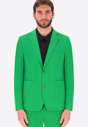 Uomo che indossa un abito verde acceso con una camicia nera con bottoni, in piedi su uno sfondo bianco uniforme, di fronte a sé.