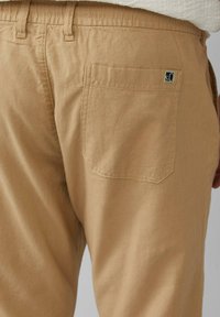 Nahaufnahme von beigen Hosen, die die Gesäßtasche mit einem kleinen dunklen Etikett in der oberen rechten Ecke zeigen.