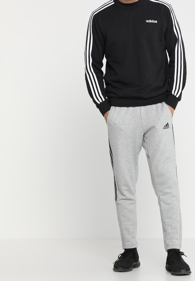 Sweat-shirt noir avec un design à trois rayures blanches sur les manches et un logo. Associé à un pantalon de survêtement gris avec des rayures noires et un logo sur le côté.
