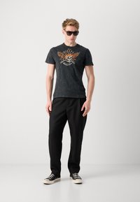 Melna grafiskā T-kreklā ar oranžu skeleta un spārnu dizainu, apvienots ar brīvi pieguļošu melnu biksēm un melniem sporta apaviem. Modelis nēsā saulesbrilles.