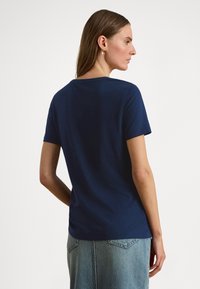 Žena s dugom smeđom kosom koja nosi zlatne naušnice u obliku obruča, mornarskoplavu majicu kratkih rukava i svijetloplavu klasičnu traper suknju, gledana s leđa.