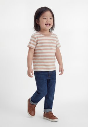 Enfant souriant marchant, portant un t-shirt rayé beige et blanc, un jean bleu foncé retroussé et des baskets marron sur fond blanc uni.