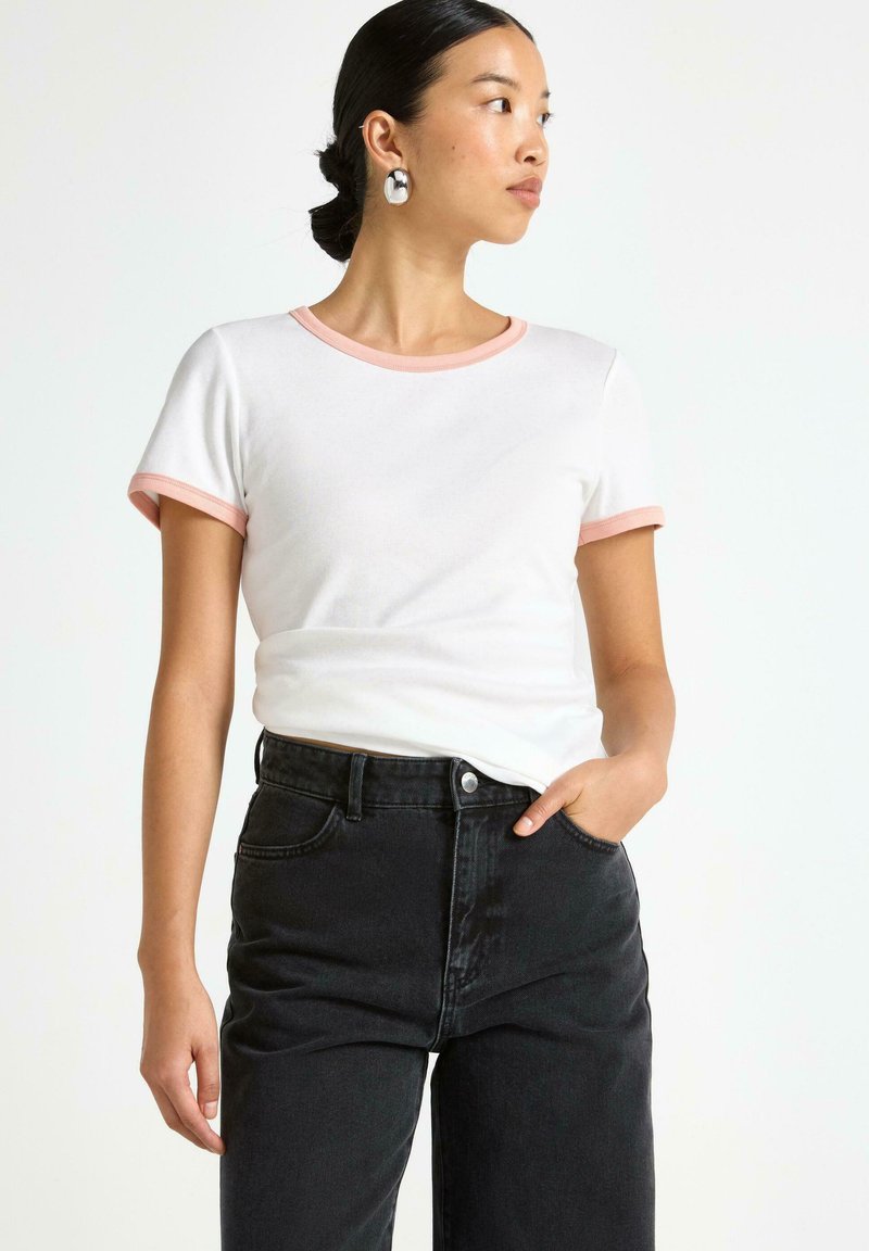 Kvinde med mørkt hår i en knold iført en hvid t-shirt med lyserøde kanter og sorte højtaljede jeans, ser mod højre med den ene hånd i lommen.