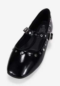 Bailarina de charol negro con punta cuadrada, adornos de tachuelas plateadas a lo largo de la correa y detalle de hebilla en el tobillo. Textura suave.