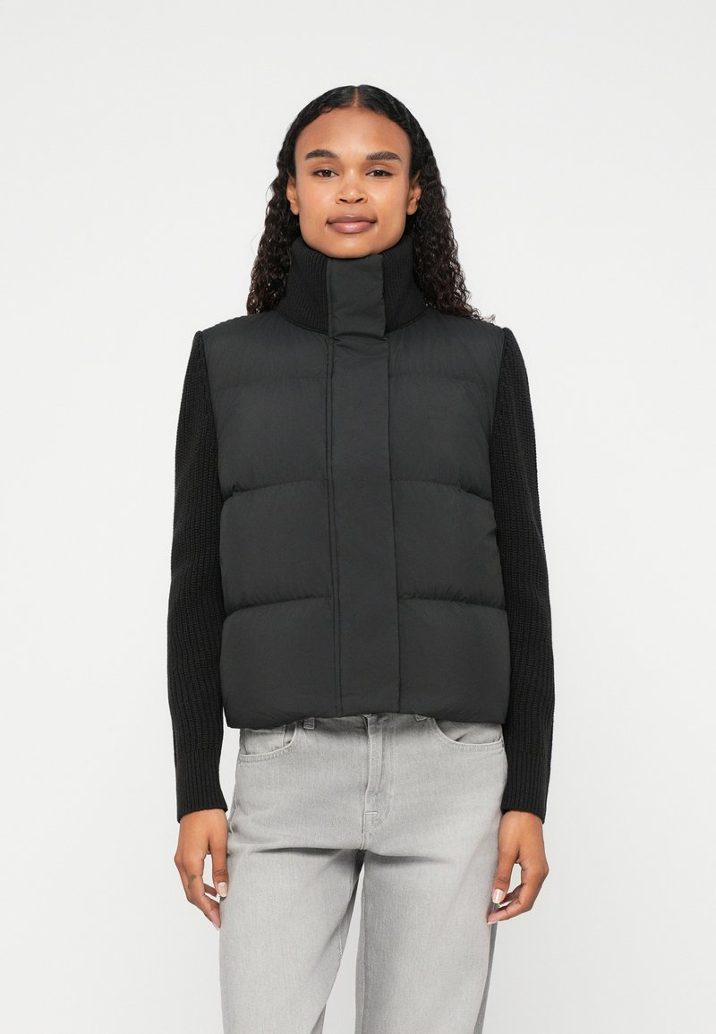 Črn napihnjen telovnik z visokim ovratnikom in prešitim dizajnom, vparen z črnim pletenim dolgim topom. Mehek material, krojena oblika. Svetlo sivi džins.