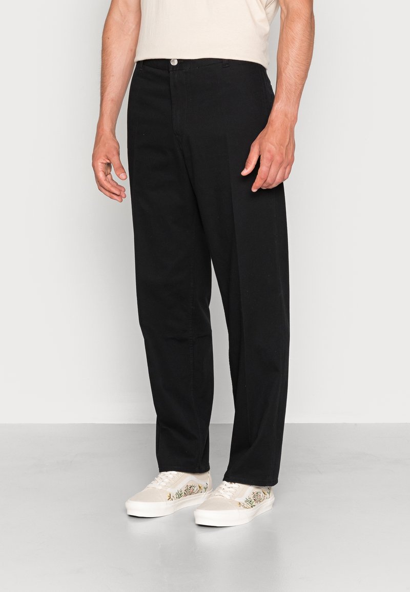 Black cotton trousers with a straight fit, front button closure, and two side pockets, paired with light-colored embroidered sneakers.