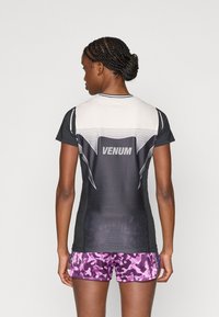 Μαύρο και γκρι κοντομάνικο πουκάμισο με διχτυωτό σχέδιο, με το λογότυπο "Venum" σε λευκό. Συνδυάζεται με μοβ σορτς καμουφλάζ.