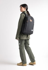 Mochila de tela negra con un acento de cuero geométrico, bolsillo lateral, tirantes ajustables, llevada por una persona con atuendo verde y calzado color arena.