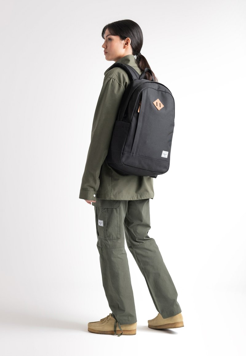 Mochila de tela negra con un acento de cuero geométrico, bolsillo lateral, tirantes ajustables, llevada por una persona con atuendo verde y calzado color arena.