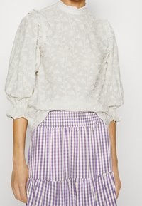 Blouse blanche brodée avec motif floral, manches bouffantes et bordure à volants, assortie à une jupe rayée violette et blanche.
