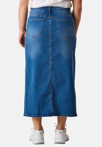 Denim skirt with a long, straight cut, in medium blue. Features two back pockets, a waistband, and a raw hem. Worn with white sneakers.