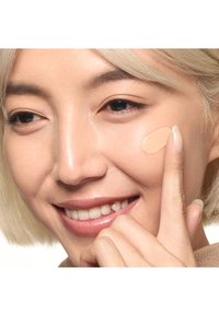 Personne souriante aux cheveux clairs appliquant un fond de teint liquide beige sur la joue avec le bout des doigts, montrant un gros plan de l'application du maquillage.