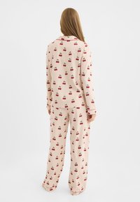 Mujer vistiendo un pijama color crema con un patrón de cerezas rojas y rayas naranja claro, de pie descalza, mirando hacia atrás sobre un fondo blanco liso.