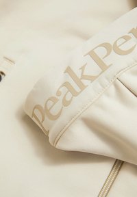 Beež jaki kude, millel on sile tekstuur, kaelarihmaga suur heleda pruuni "Peak Performance" logo ja tõmblukk.