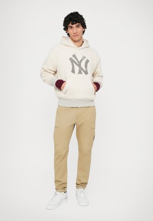 Hanorac bej cu un logo gri, manșete bordeau și buzunare. Asortat cu pantaloni cargo bej și adidași albi. Textură de fabrică netedă.