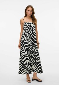 Robe maxi rayée noir et blanc avec de fines bretelles spaghetti, présentant une coupe ample et un tissu texturé. Des sandales plates complètent le look.