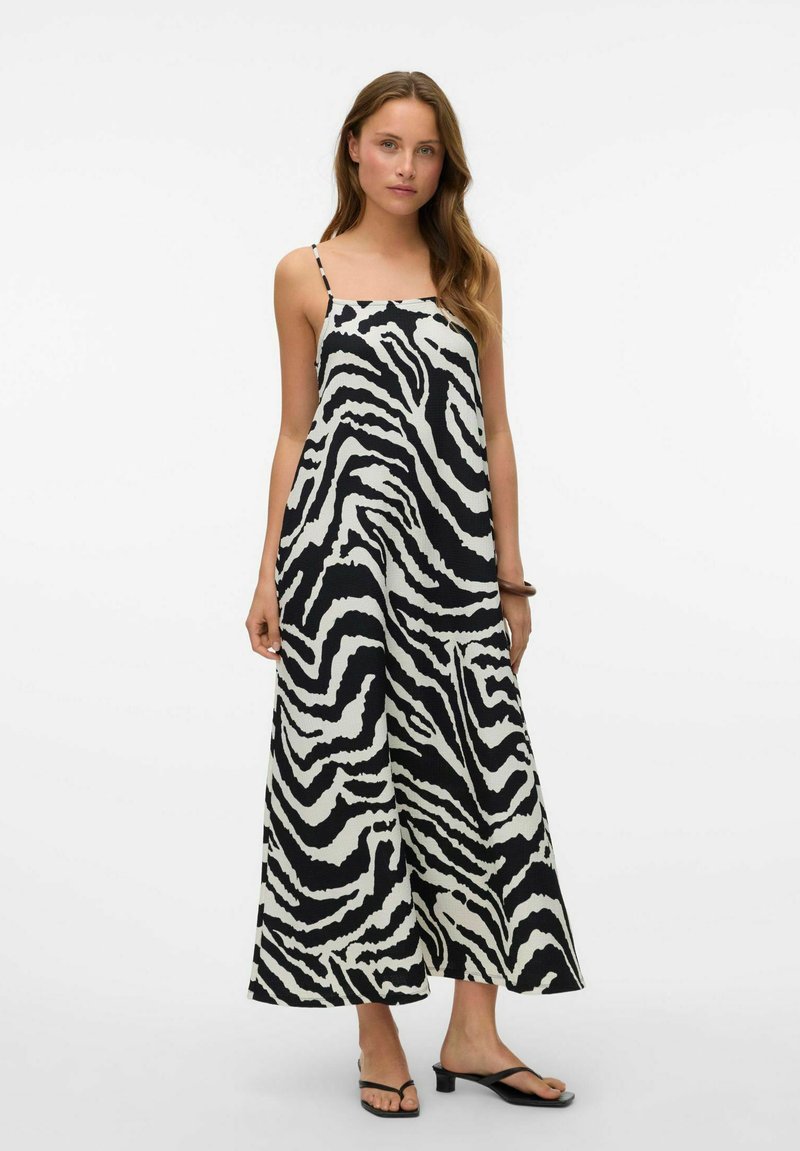 Robe maxi rayée noir et blanc avec de fines bretelles spaghetti, présentant une coupe ample et un tissu texturé. Des sandales plates complètent le look.