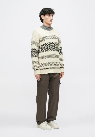 Beige kötött pulóver fekete geometrikus mintákkal, barna cargo nadrággal és fehér sportcipővel párosítva. A modell kezeit a zsebében tartja.