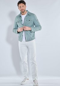 Hellpetrolfarbene Hemdjacke mit zwei Brusttaschen, getragen über einem weißen T-Shirt, kombiniert mit weißen Jeans und weißen Turnschuhen. Glatte Textur.