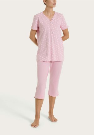 Donna che indossa un top rosa chiaro a maniche corte con motivo floreale bianco e pantaloni corti coordinati, in piedi a piedi nudi su sfondo chiaro.