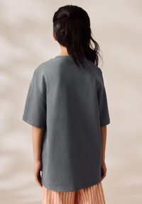 Person with dark hair tied back wears oversized gray t-shirt and striped pink, yellow, and orange pants standing against plain background.