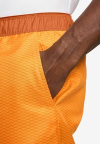 Orangene Shorts mit einem Diamantmuster, ausgestattet mit einem braunen Bund und einer Tasche. Das Material ist leicht und glatt.