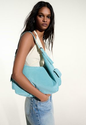 Mladá žena s dlhými tmavými vlasmi, oblečená v bielom tielku bez rukávov a džínsach, nesie veľkú svetlomodrú semišovú kabelku cez rameno.