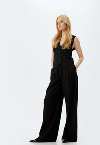 Schwarze Weste mit fünf Knöpfen, kombiniert mit weit geschnittenen schwarzen Hosen. Glattes Material mit minimalen Details und einem maßgeschneiderten Schnitt.