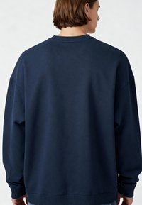 Tamsiai mėlynas per didelis džemperis, tekstūruota medžiaga, banguota apykaklė ir rankogaliai, kritimo pečių dizainas, minimalus siuvinėjimo detalės, be grafikos.