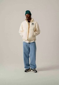 White puffer jacket with an emblem, paired with light blue jeans. The model wears a green knitted beanie and black sneakers.