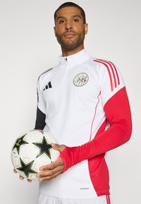 Hvid atletisk top med røde og sorte detaljer, med en lynlåskrave og logo. Holder en mønstrede fodbold med sorte stjerner.
