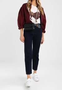 Donna che indossa una giacca cropped borgogna, una maglietta bianca con stampa di leone, jeans scuri, sneakers bianche e una borsa a tracolla nera trapuntata.