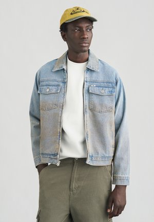 Lyseblå denimjakke med krave og to brystlommer. Bæres over en hvid skjorte, kombineret med grønne cargobukser.