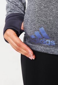 Ręka ciągnąca za boczną kieszeń szarej kurtki Adidas z czarnymi mankietami, noszonej na czarnych spodniach.