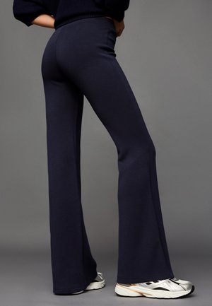 Person trägt marineblaue Schlaghosen und silberweiße Turnschuhe und steht vor einem schlichten grauen Hintergrund.