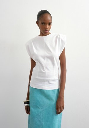 Frau trägt ein weißes, gepolstertes Oberteil mit Schulterpolstern und einen türkisfarbenen Rock, steht vor einem schlichten weißen Hintergrund und hält Armbänder an ihrem linken Handgelenk.