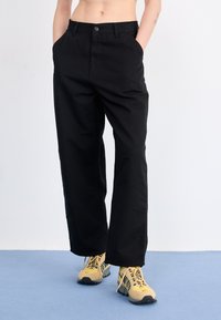 Pantalon en coton noir avec une coupe droite, doté de poches avant et d'une fermeture à bouton unique. Porté avec des baskets jaunes et noires.