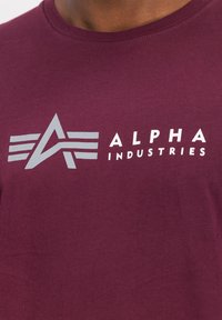 Bordeaux bomulds T-shirt med et sølv logo med stiliserede bogstaver og vinger. Rund halsudskæring og korte ærmer.