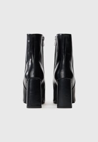 Par de botas de cuero negras hasta el tobillo con tacones anchos y cremalleras traseras, mostradas desde la parte trasera sobre un fondo blanco liso.