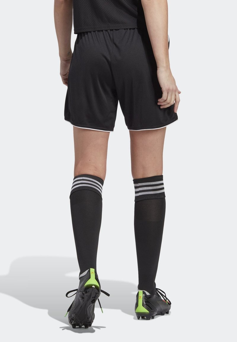 Personne portant un short de football noir, des chaussettes noires montantes avec des rayures blanches et des chaussures à crampons noires avec des touches vertes, vue de dos.