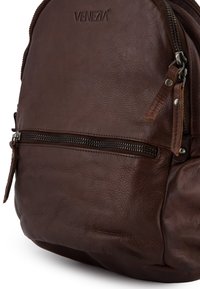 Brauner Leder-Rucksack mit strukturierter Oberfläche, ausgestattet mit einer vorderen Reißverschlusstasche und einem Markenlogo. Kompakte Form mit detaillierter Naht.