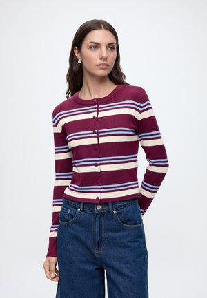 Vrouw met lang donker haar die een bordeauxrood, wit en blauw gestreept, geknoopt vest en blauwe spijkerbroek draagt tegen een effen lichte achtergrond.