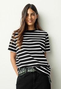 Črno-bela majica s kratkimi rokavi in okroglim izrezom. Oblečena s temnimi hlačami in pasom z vzorcem. Gladek teksturiran material.